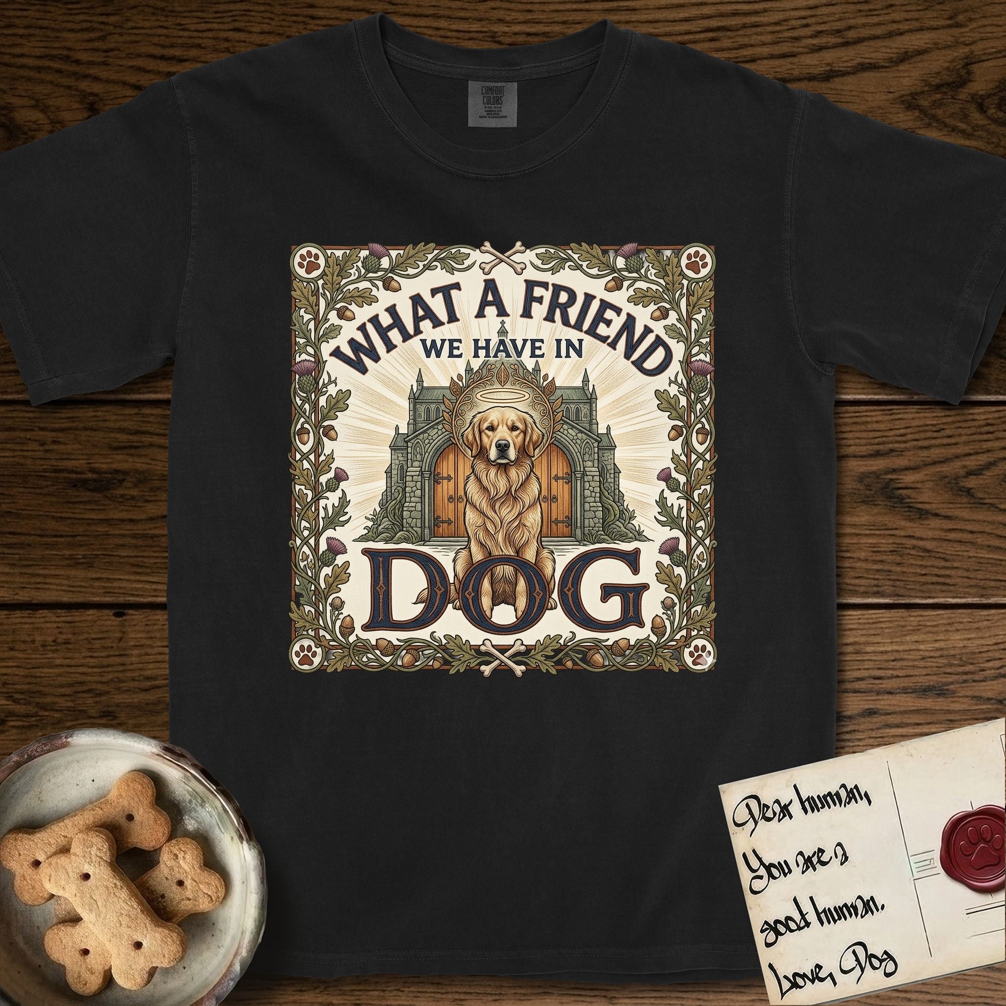 What a Friend We Have in Dog T-Shirt flat lay in Black, featuring an Arts and Crafts tapestry illustration of a haloed Golden Retriever seated before a monastery, framed by an oak-leaf, acorn, and thistle border, with the word DOG below.