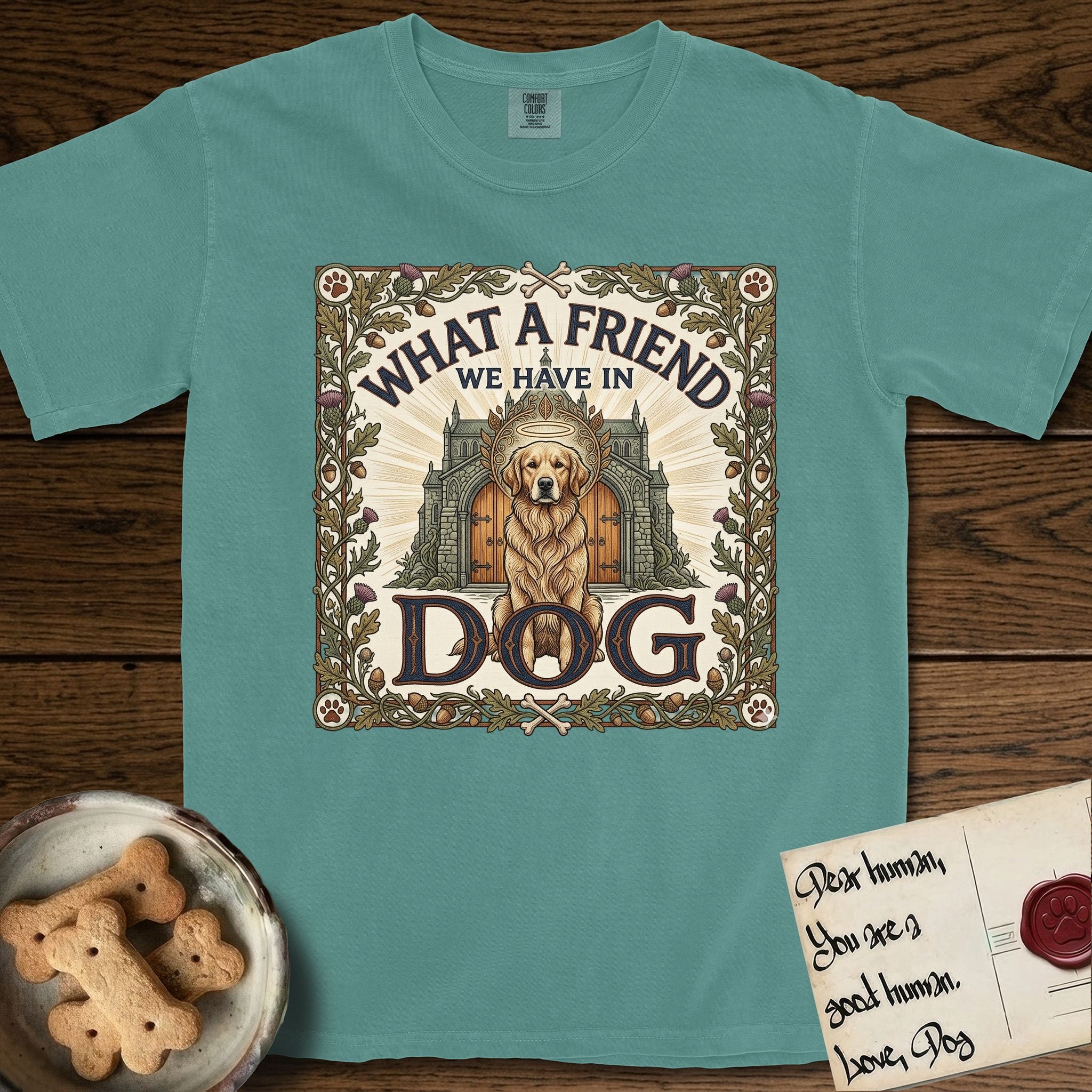 What a Friend We Have in Dog T-Shirt flat lay in Blue Spruce, featuring an Arts and Crafts tapestry illustration of a haloed Golden Retriever seated before a monastery, framed by an oak-leaf, acorn, and thistle border, with the word DOG below.
