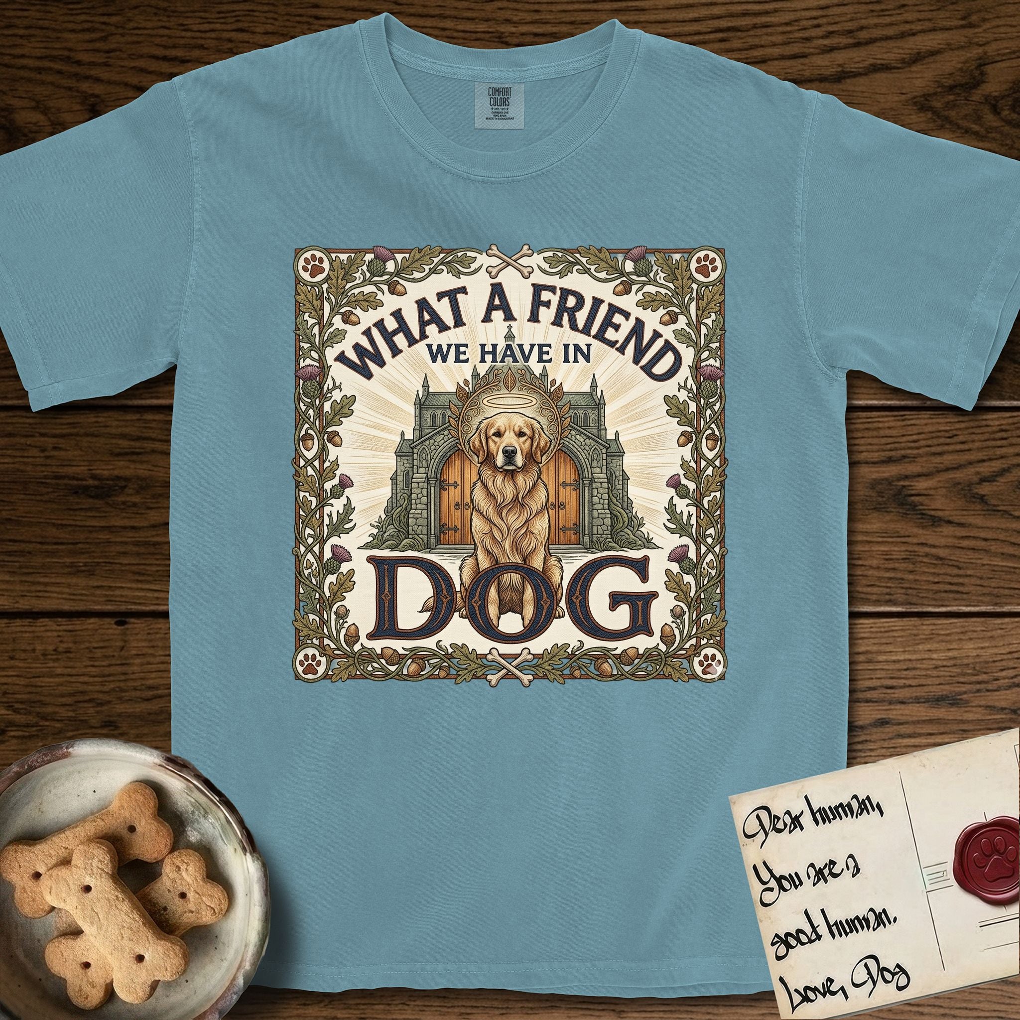 What a Friend We Have in Dog T-Shirt flat lay in Ice Blue, featuring an Arts and Crafts tapestry illustration of a haloed Golden Retriever seated before a monastery, framed by an oak-leaf, acorn, and thistle border, with the word DOG below.