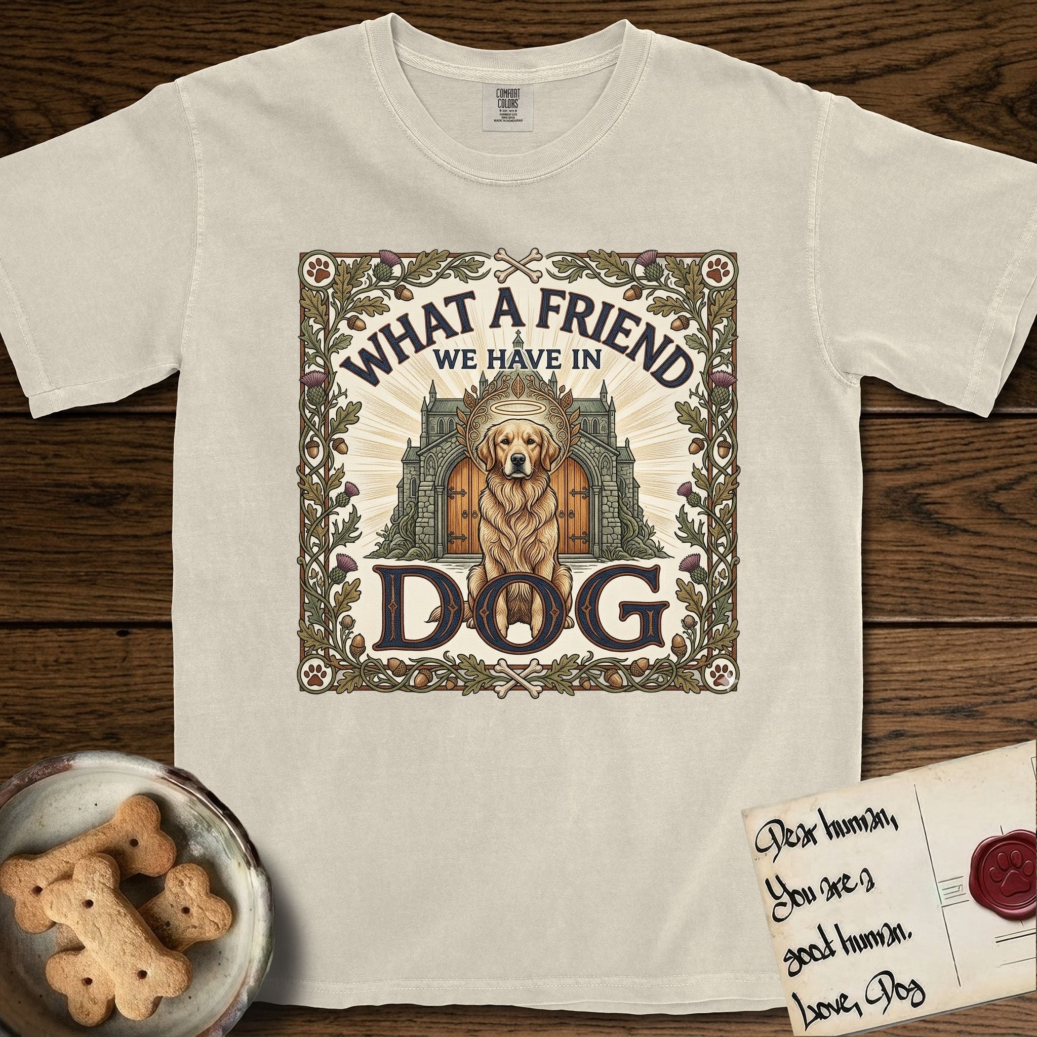 What a Friend We Have in Dog T-Shirt flat lay in Ivory, featuring an Arts and Crafts tapestry illustration of a haloed Golden Retriever seated before a monastery, framed by an oak-leaf, acorn, and thistle border, with the word DOG below.