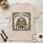 What a Friend We Have in Dog T-Shirt flat lay in Sand, featuring an Arts and Crafts tapestry illustration of a haloed Golden Retriever seated before a monastery, framed by an oak-leaf, acorn, and thistle border, with the word DOG below.
