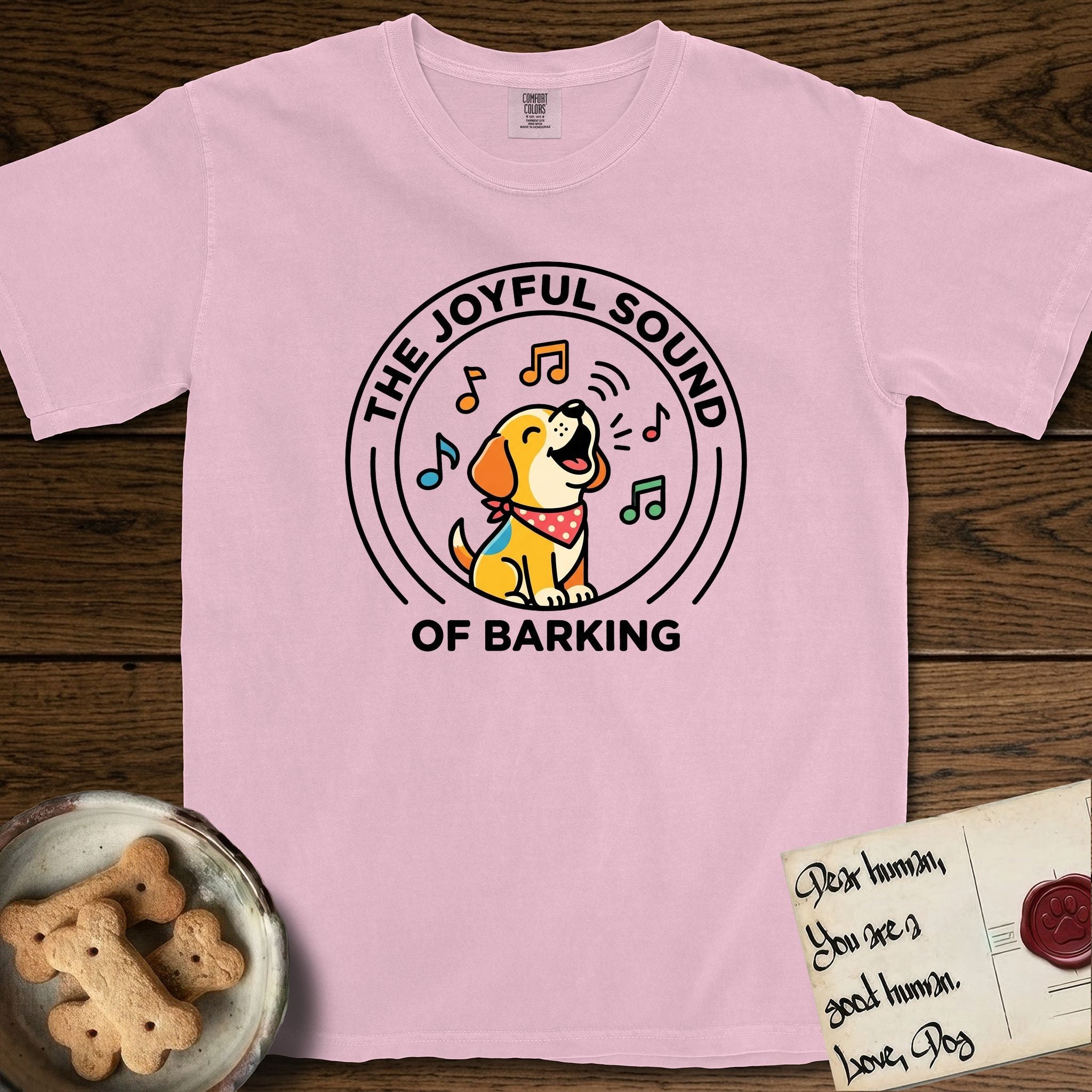 The Joyful Sound of Barking T-Shirt flat lay in Blossom, featuring a flat vector cartoon emblem of a happy puppy with lifted ears surrounded by musical notes, ringed by the words The Joyful Sound of Barking.