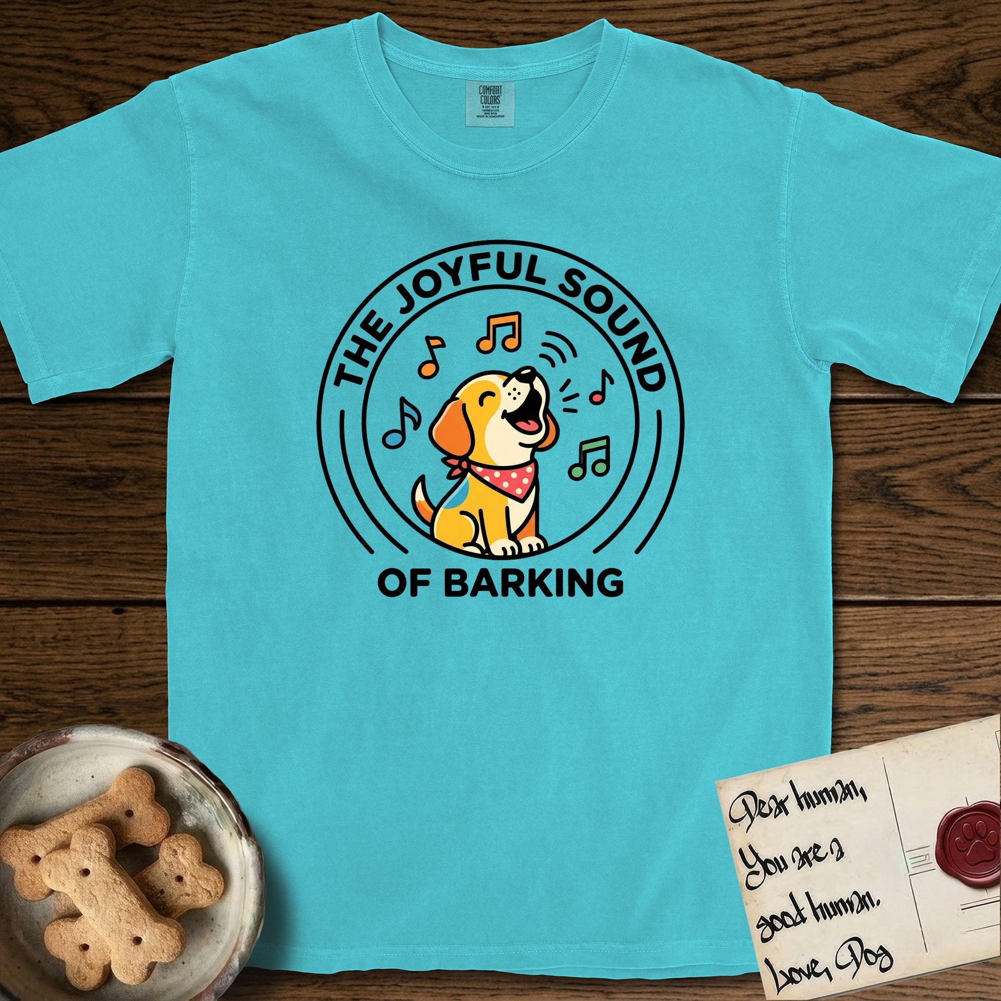 The Joyful Sound of Barking T-Shirt flat lay in Lagoon Blue, featuring a flat vector cartoon emblem of a happy puppy with lifted ears surrounded by musical notes, ringed by the words The Joyful Sound of Barking.