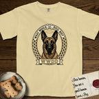What Would Dog Do Belgian Malinois T-Shirt flat lay in Butter, featuring a vintage gold portrait medallion of a haloed Belgian Malinois in an ornamental rosette-scroll border with the question What Would Dog Do.