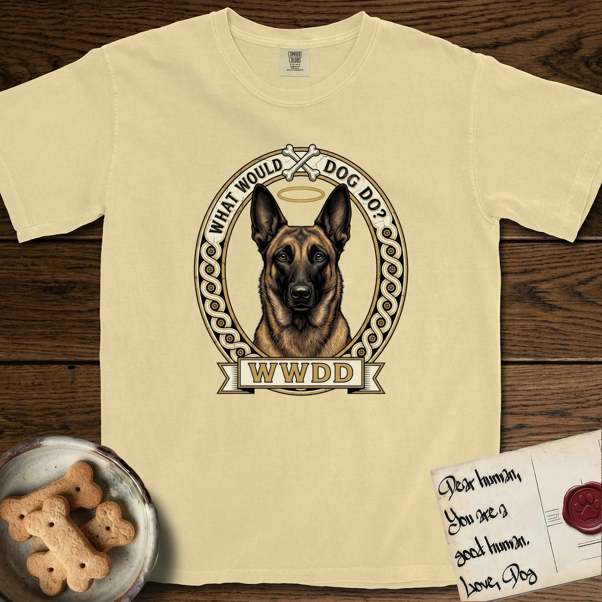 What Would Dog Do Belgian Malinois T-Shirt flat lay in Butter, featuring a vintage gold portrait medallion of a haloed Belgian Malinois in an ornamental rosette-scroll border with the question What Would Dog Do.