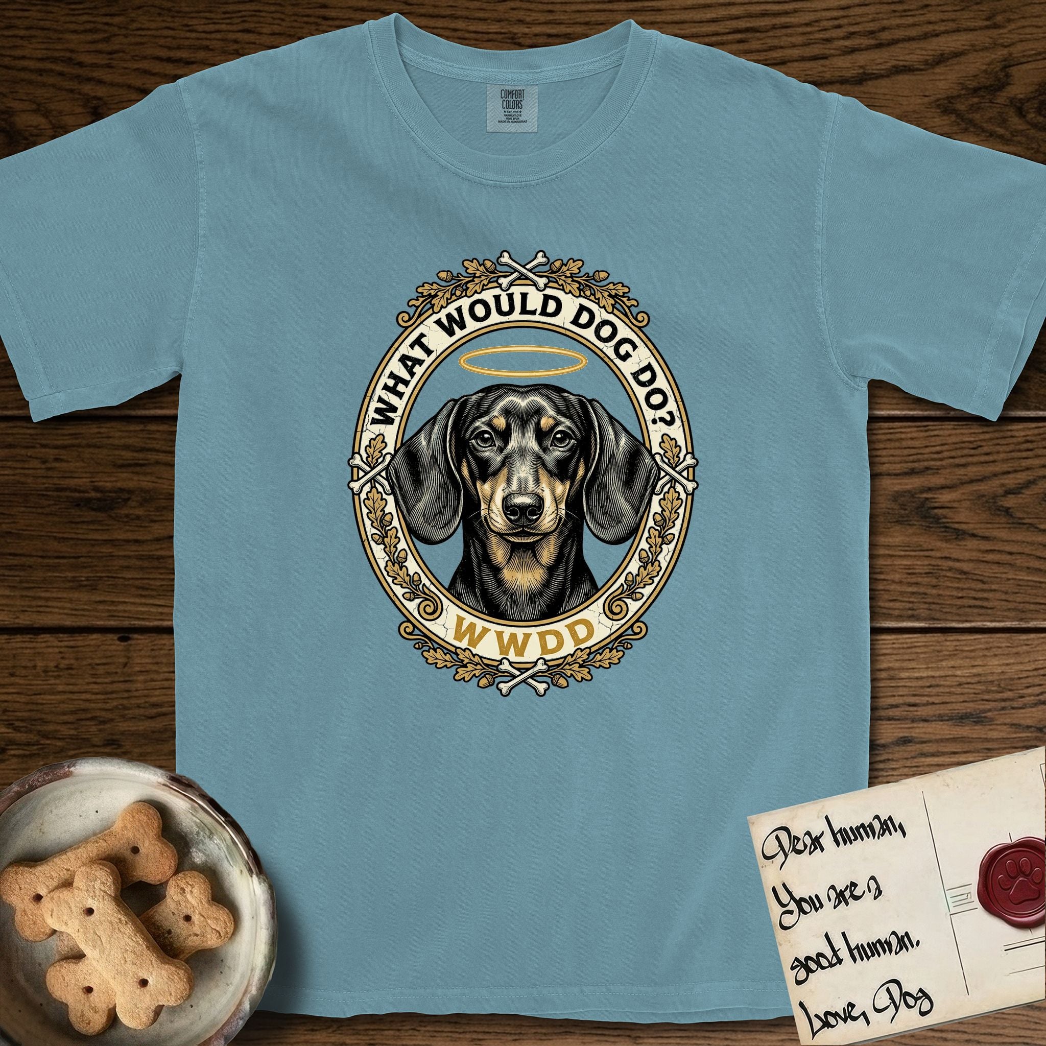 What Would Dog Do Dachshund T-Shirt flat lay in Ice Blue, featuring an oak-leaf and acorn framed gold portrait medallion of a haloed Dachshund with the question What Would Dog Do.