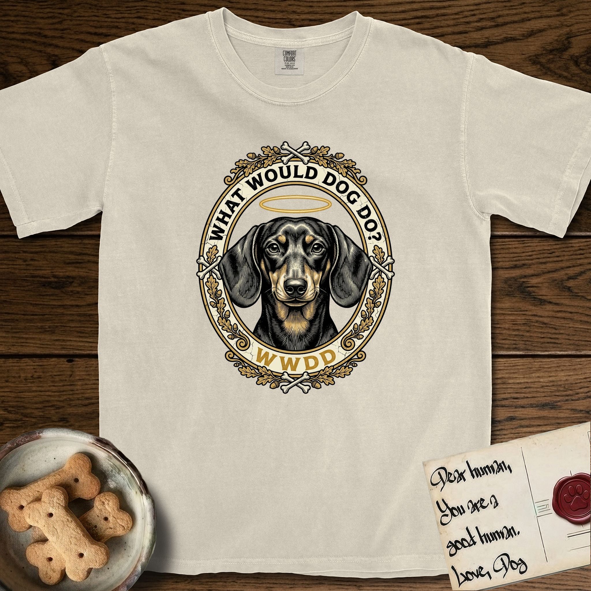 What Would Dog Do Dachshund T-Shirt flat lay in Ivory, featuring an oak-leaf and acorn framed gold portrait medallion of a haloed Dachshund with the question What Would Dog Do.