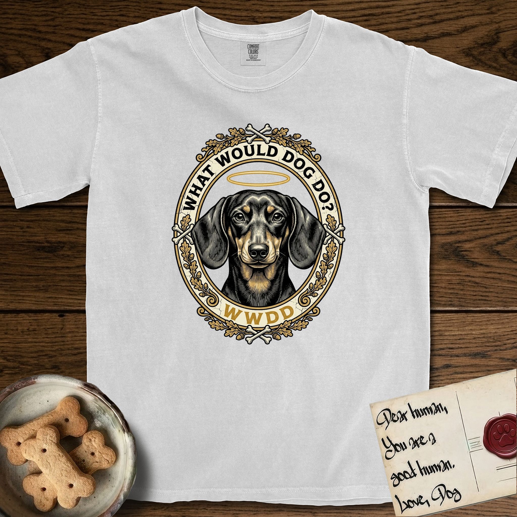 What Would Dog Do Dachshund T-Shirt flat lay in White, featuring an oak-leaf and acorn framed gold portrait medallion of a haloed Dachshund with the question What Would Dog Do.
