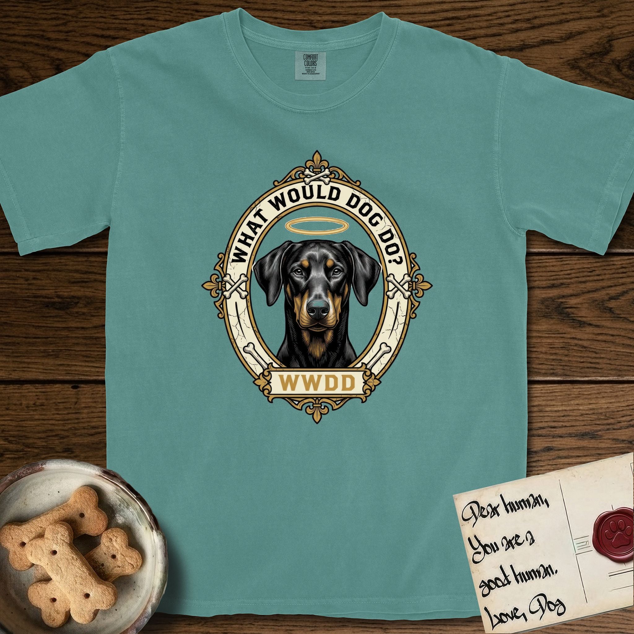 What Would Dog Do Doberman T-Shirt flat lay in Blue Spruce, featuring a fleur-de-lis framed gold portrait medallion of a haloed Doberman with natural uncropped ears and the question What Would Dog Do.
