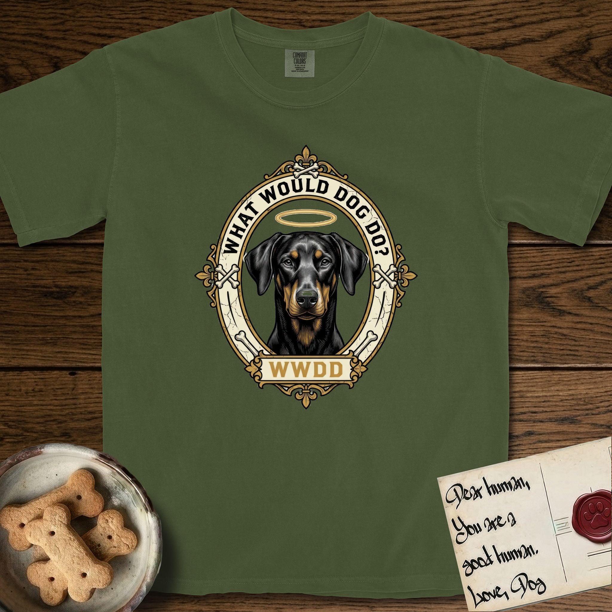 What Would Dog Do Doberman T-Shirt flat lay in Hemp, featuring a fleur-de-lis framed gold portrait medallion of a haloed Doberman with natural uncropped ears and the question What Would Dog Do.