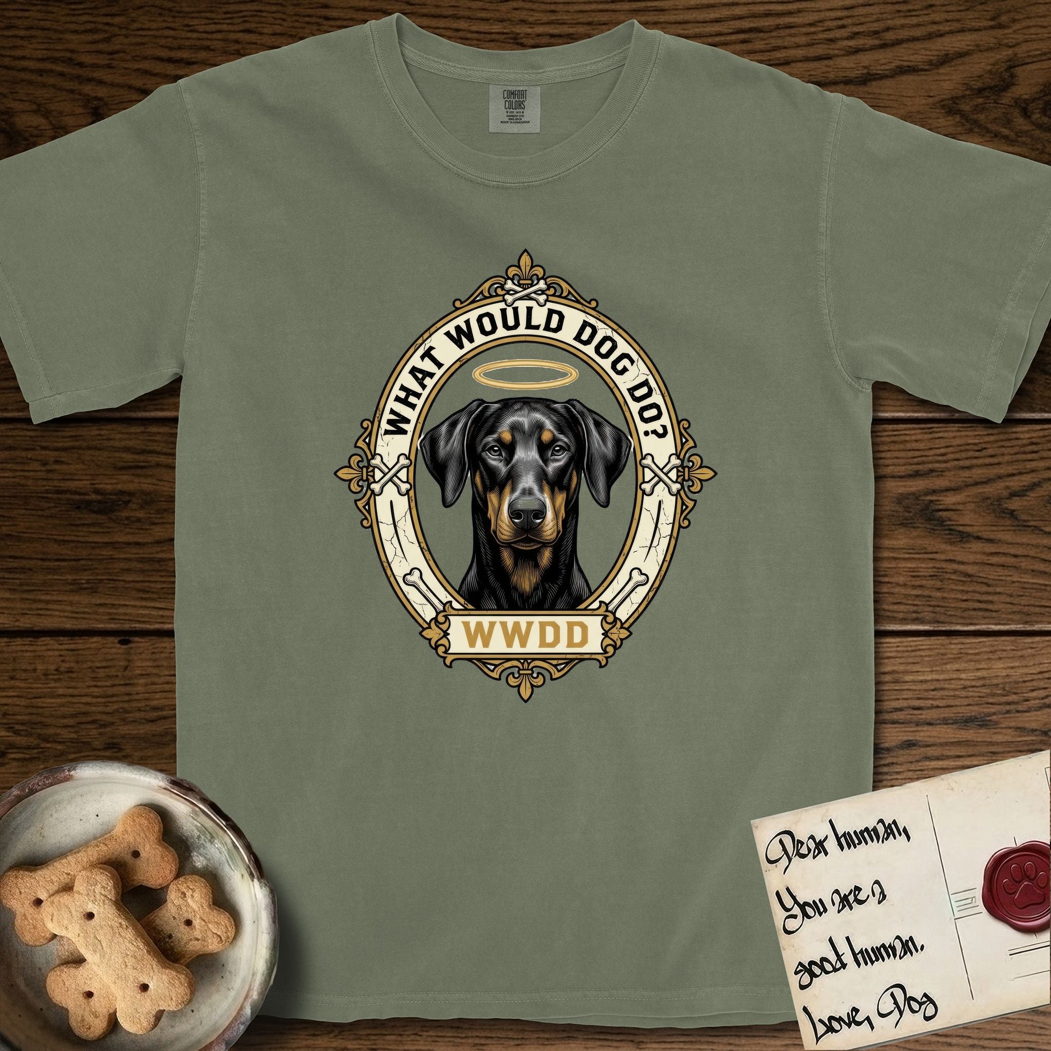 What Would Dog Do Doberman T-Shirt flat lay in Sage, featuring a fleur-de-lis framed gold portrait medallion of a haloed Doberman with natural uncropped ears and the question What Would Dog Do.