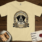 What Would Dog Do Saint Bernard T-Shirt flat lay in Butter, featuring a vintage gold portrait medallion of a haloed Saint Bernard in an alpine laurel wreath with edelweiss blossoms and the question What Would Dog Do.