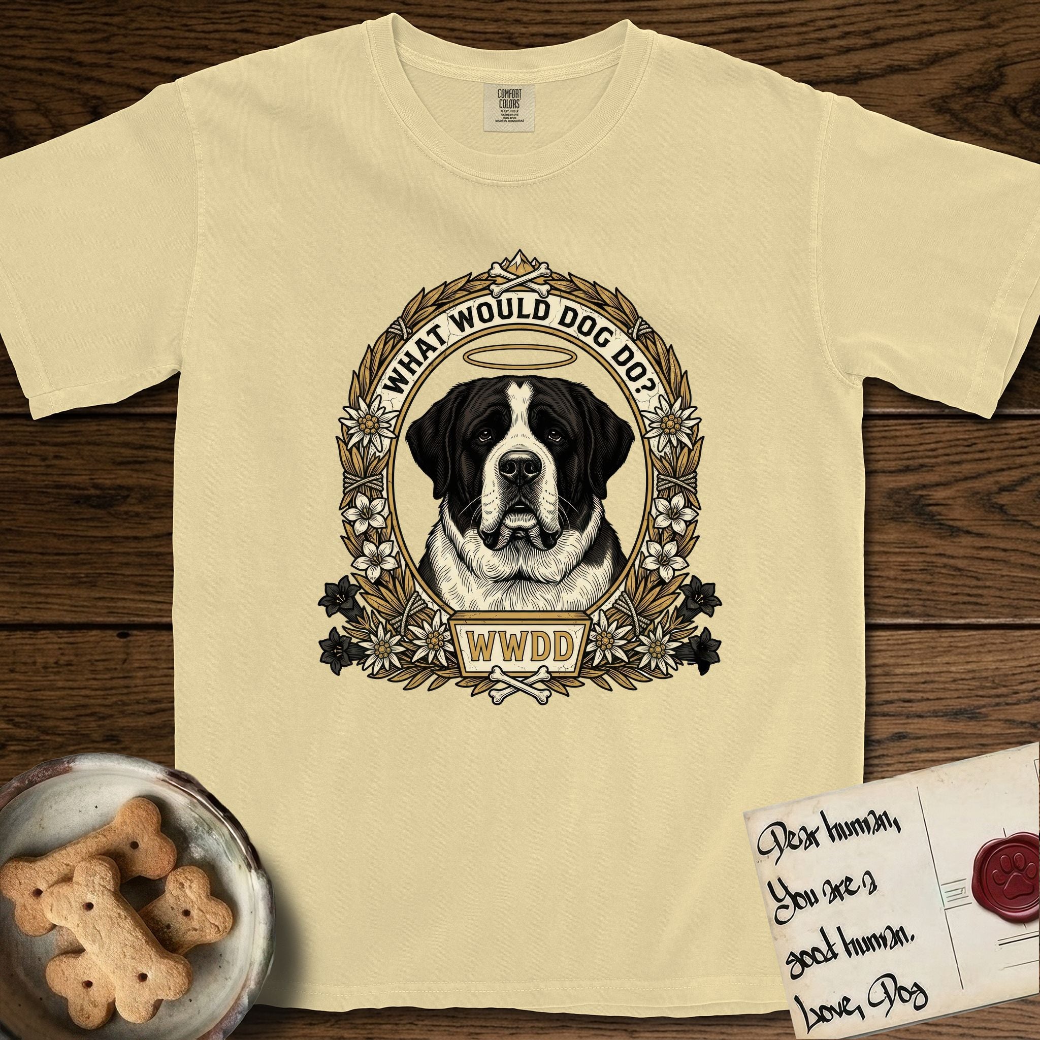 What Would Dog Do Saint Bernard T-Shirt flat lay in Butter, featuring a vintage gold portrait medallion of a haloed Saint Bernard in an alpine laurel wreath with edelweiss blossoms and the question What Would Dog Do.