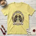 What Would Dog Do Spaniel T-Shirt flat lay in Cornsilk, featuring a rose-bordered gold portrait medallion of a haloed Cavalier King Charles Spaniel with the question What Would Dog Do.