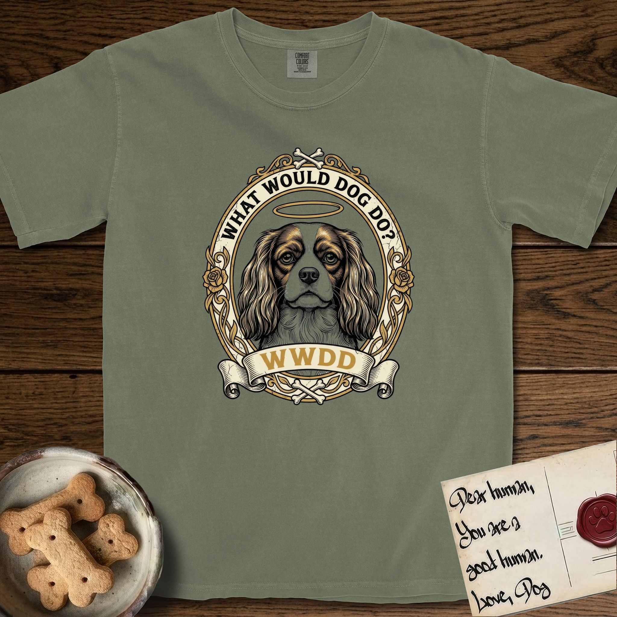 What Would Dog Do Cavalier T-Shirt flat lay in Sage, featuring a rose-bordered gold portrait medallion of a haloed Cavalier King Charles Spaniel with the question What Would Dog Do.