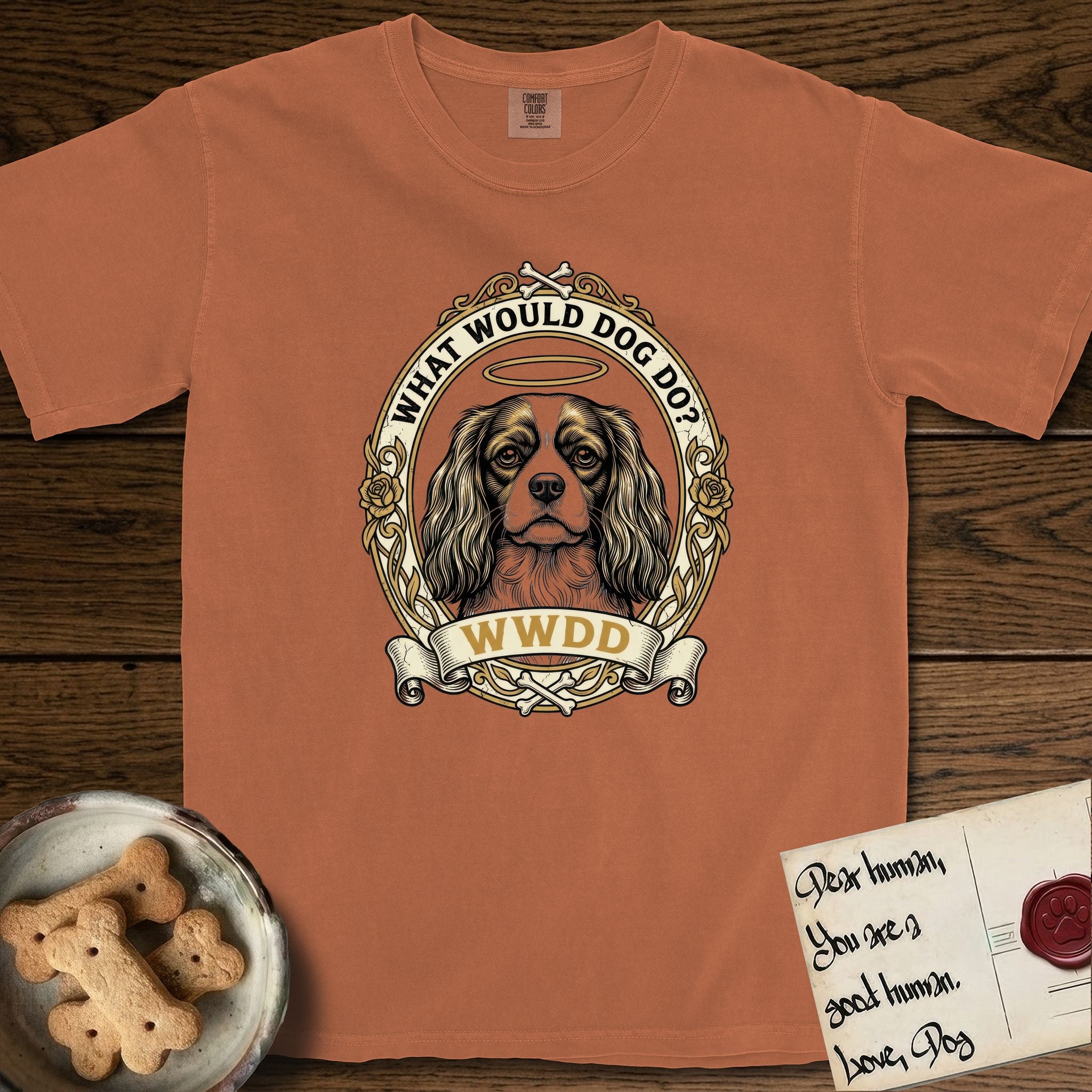 What Would Dog Do Cavalier T-Shirt flat lay in Yam, featuring a rose-bordered gold portrait medallion of a haloed Cavalier King Charles Spaniel with the question What Would Dog Do.
